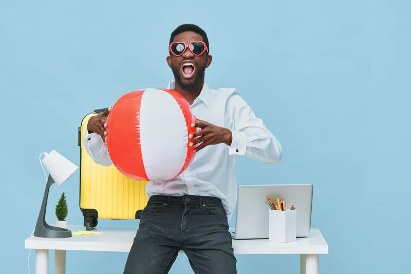 Energetic man in sunglasses holding a large beach ball smiling happily in a modern office setting with bright blue background, casual white shirt, and dark jeans. People lifestyle concept, summer vaca