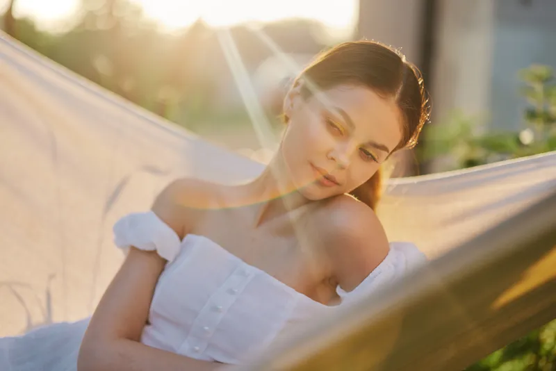 Young woman in a white dress relaxing in a hammock at sunset, symbolizing tranquility and summer vibes Natural light and soft colors create a serene atmosphere