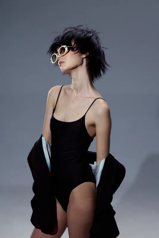 Stylish woman in black bodysuit and sunglasses posing with confident expression against gray background