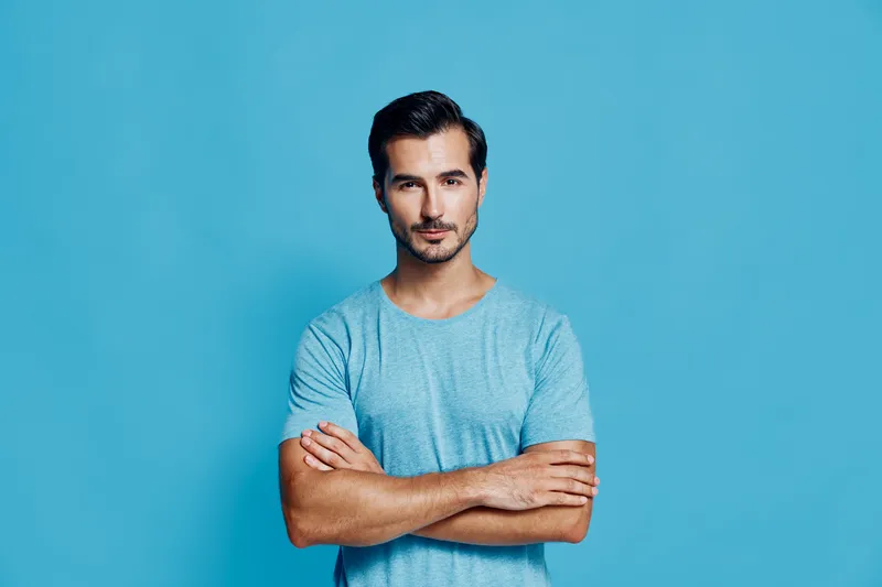Confident man standing with crossed arms, wearing a casual blue t-shirt, looking directly at the camera against a solid blue background. Professional style concept.