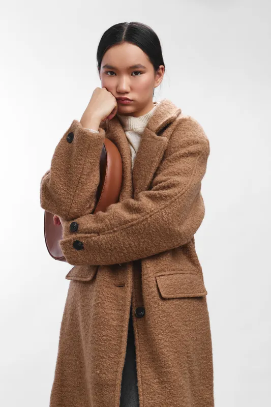 asian woman wearing a brown coat stands in a minimal studio setting, carrying a backpack and a thoughtful expression, creating a calm modern fashion mood and urban vibe.