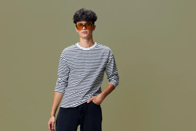 young man in striped shirt wearing sunglasses stands confidently with hand in pocket against simple beige background, casual style, modern fashion look
