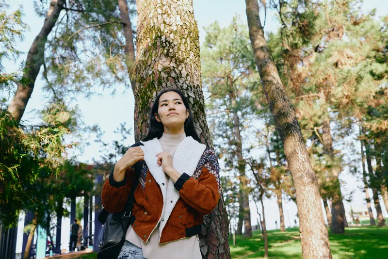 Woman leaning against a tree in a sunlit park, wearing a warm jacket and scarf, enjoying nature among tall pines for a calm outdoor moment blending fashion and forest atmosphere