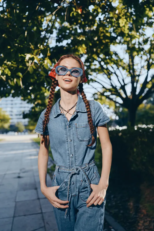 lifestyle woman wearing modern boho-western denim jumpsuit and sunglasses outdoors, vibrant film color street style with natural sunlight and greenery background