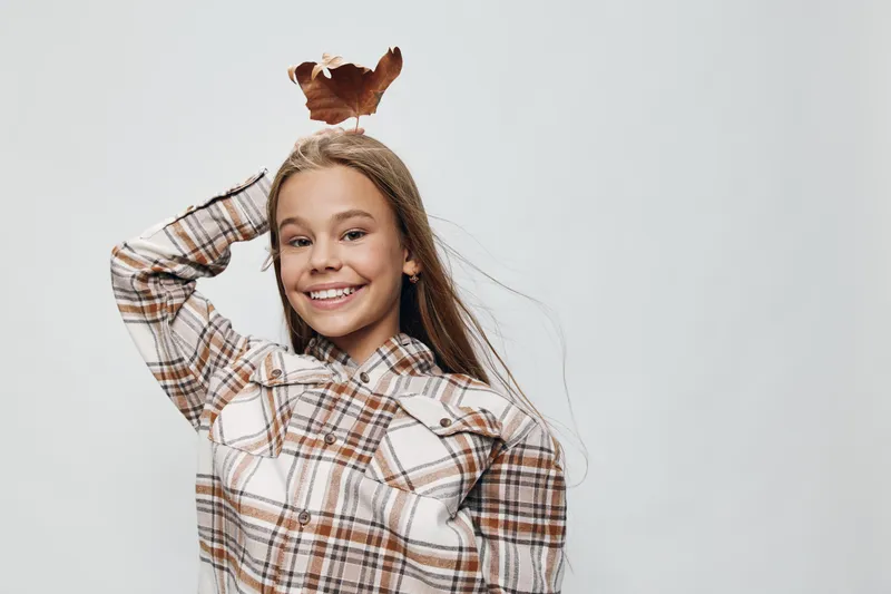 Smiling girl with long hair wearing plaid shirt holding autumn leaf on head, posing against plain background with happy playful expression in studio indoors.