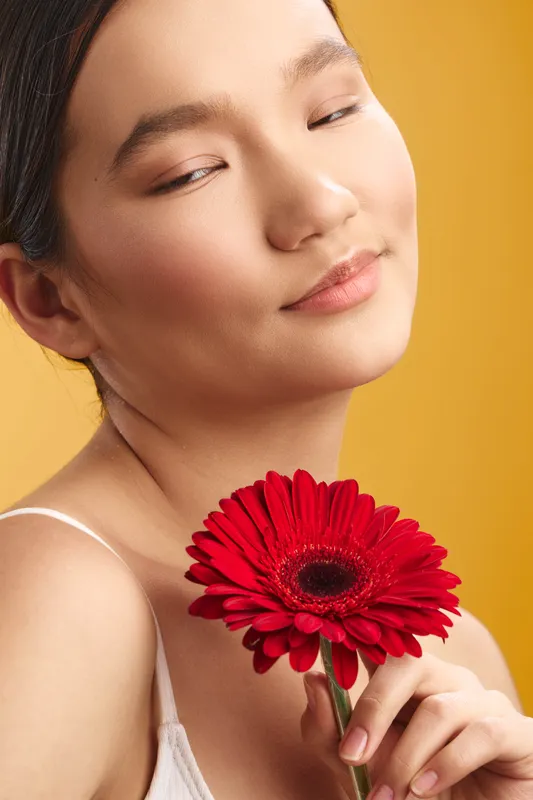 asian woman with a serene smile holds a vivid red gerbera near her face, soft focus background in warm orange, creating a calm, elegant closeup portrait.