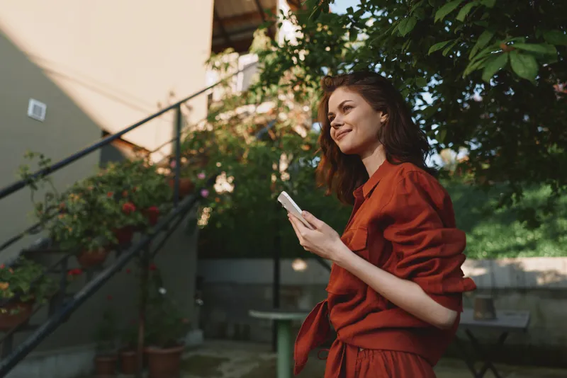 An outdoor lifestyle scene shows a woman in a red dress smiling while holding a phone, standing in a sunny garden. Concept for casual living and everyday moments and memories