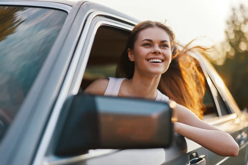 Smiling young woman enjoying a road trip with her hair blowing in the wind, framed by the car window in a natural setting at sunset.