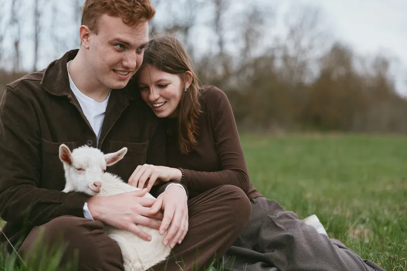 A young couple sits on grass in casual clothes, gently holding a white baby goat while sharing a tender moment outdoors in a natural lifestyle setting during early spring.