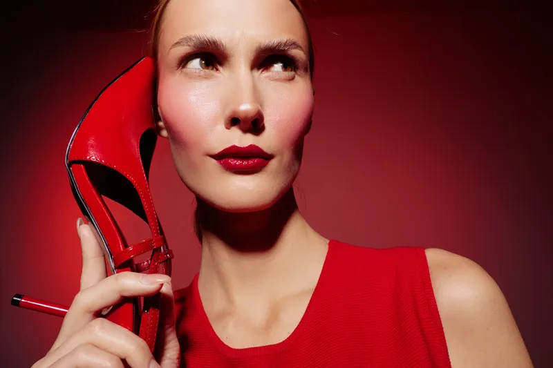 Woman in red dress holds red high heel shoe near face with confident expression. Studio portrait with red background. Concept of fashion, style, and elegance.