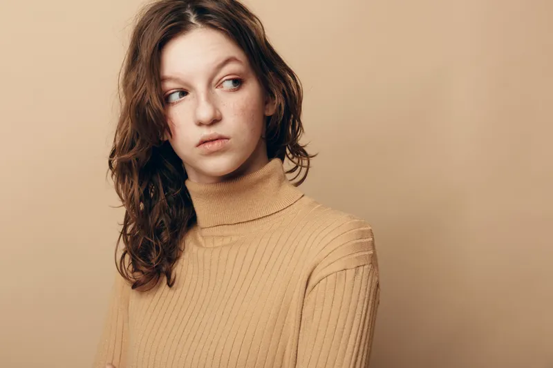 Young woman with curly hair in beige sweater looks away thoughtfully on beige background. Concept of contemplation, mood, and natural beauty in casual style.
