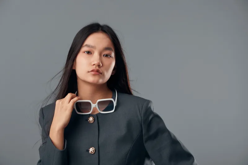 Young woman with long black hair wearing stylish gray jacket holding white sunglasses posing confidently on gray background in studio for fashion and beauty concept.