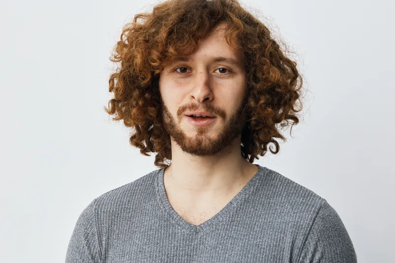 Young man with curly hair expressing confidence in a casual gray sweater, standing against a minimalistic white background, emphasizing individuality and modern style