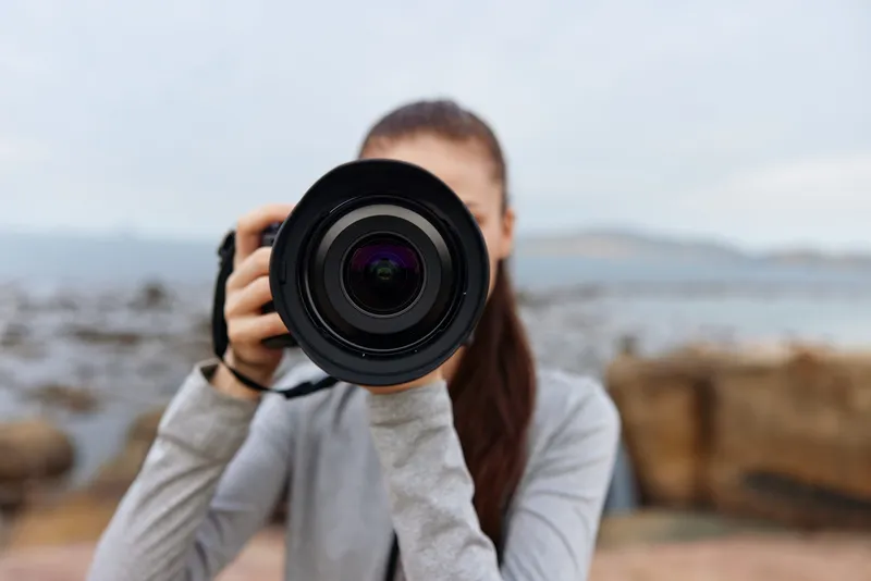Young female photographer capturing nature, holding a camera, wearing casual gray outfit, with a serene coastal background and soft, muted colors