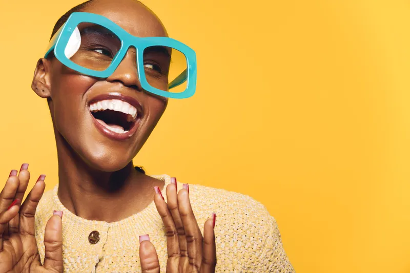 Smiling black woman with dark skin wearing oversized blue glasses and beige sweater laughs joyfully on yellow background. Concept of happiness, fashion, and positive emotions.