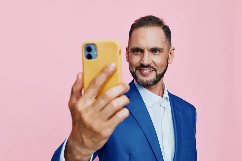 A professional man in a blue suit takes a confident selfie against a solid pink background, capturing focus and optimism in a modern business moment for branding.