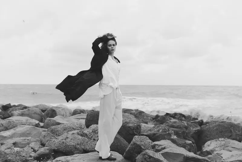 Wind swept woman on rocky coast by the sea, wearing white outfit and scarf, standing with poise as waves crash, black and white photography highlighting fashion and natural resilience