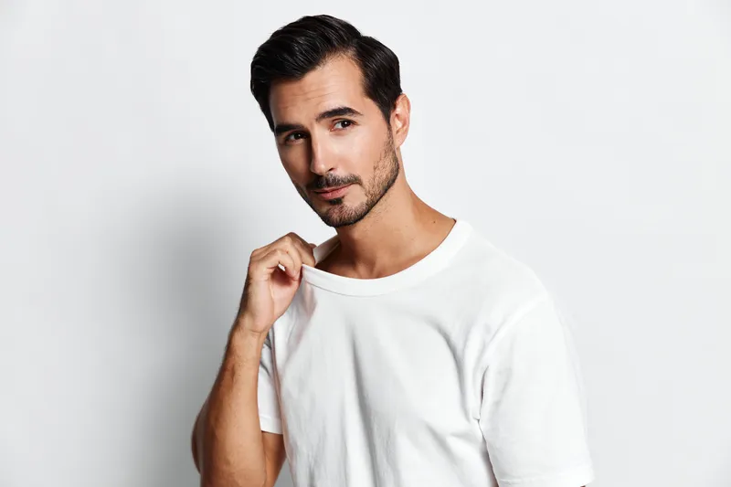Handsome young man wearing a white t-shirt with a relaxed pose, looking directly at the camera while gently tugging at his shirt collar against a minimalist background.