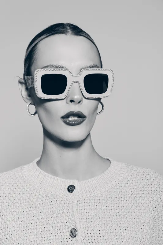 Caucasian woman with pale skin wearing large square sunglasses and hoop earrings poses in white knitted sweater on light background. Concept of fashion and accessories.
