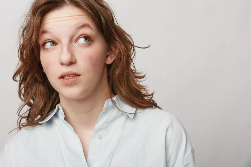 Young woman with curly hair wearing white shirt looks thoughtfully to the side on light background. Concept of contemplation, decision making, and casual lifestyle.