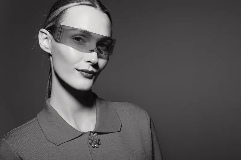 Young Caucasian woman with fair skin wearing futuristic sunglasses and a textured collar sweater, posing confidently against a dark studio background. Concept for fashion and style.