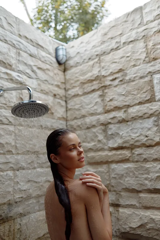 young woman taking outdoor shower with wet braided hair and natural stone wall background, calm expression in natural light, wellness concept