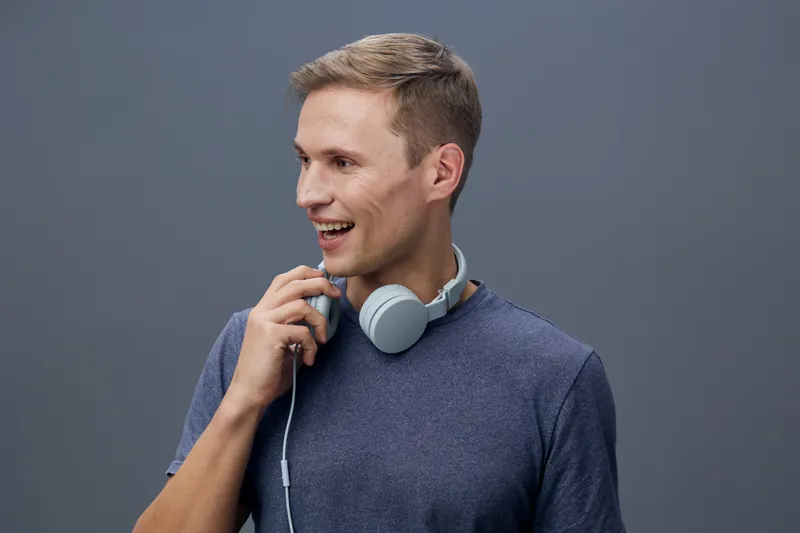 Smiling young man wearing casual blue sweater with headphones around neck, looking to side, standing against plain gray background. People lifestyle concept