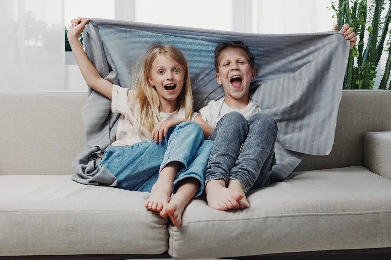 Happy playful children sitting on couch holding gray blanket behind heads, laughing and enjoying cozy home moment together in casual clothes.