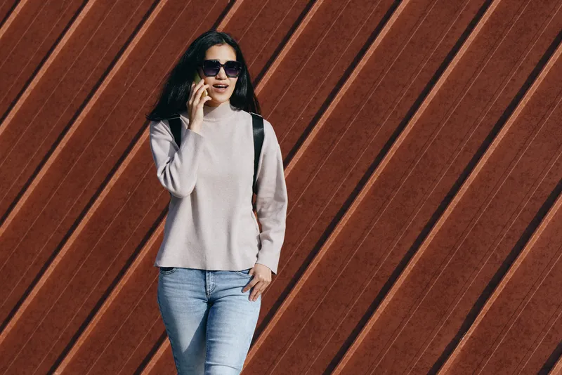 Woman in light pink sweater and jeans talks on phone, wearing sunglasses with a backpack, posed against a diagonal wooden wall for a casual street fashion moment outdoors