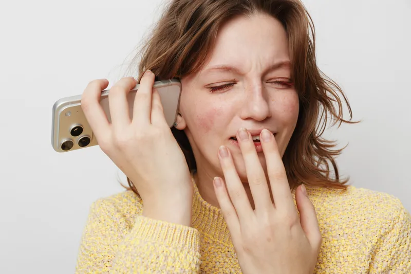 Young woman in yellow sweater holds smartphone near ear with eyes closed and hand covering mouth. Concept of emotional reaction, surprise, and communication on white background.