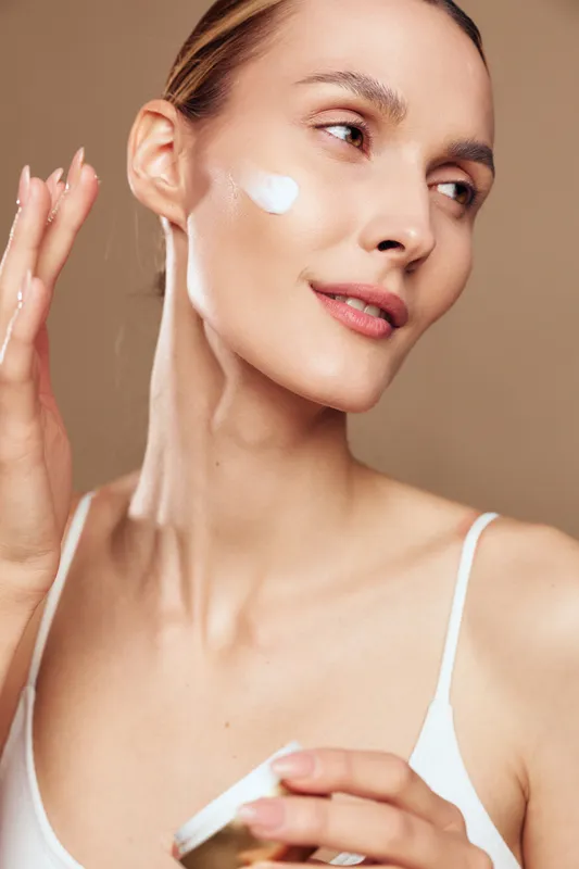 Young woman skincare closeup applying cream on face, white cotton pad and moisturizer on cheek, gentle beauty routine, spa hygiene, serene expression, beige studio background