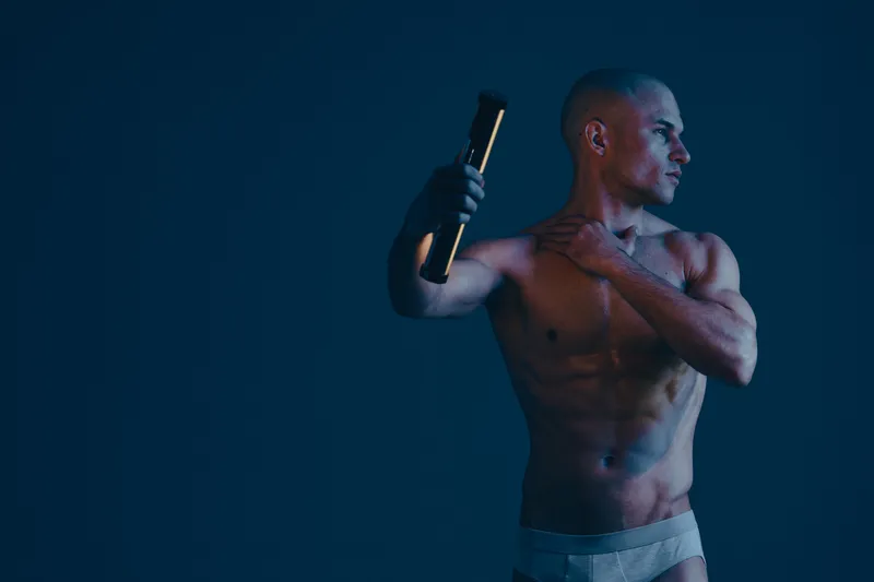 Athletic man posing with a flashlight, showcasing his toned physique against a dark blue background. Captivating blend of light and shadows enhances the visual appeal.
