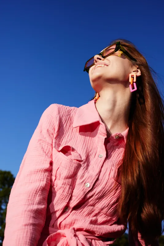Stylish young woman in a pink shirt dress, enjoying the sunshine with oversized sunglasses, showcasing a cheerful emotion against a clear blue sky
