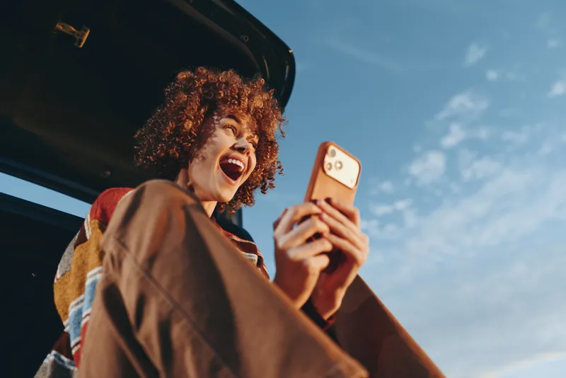 Woman lifestyle selfie, curly hair smiling outdoors near car, holding smartphone in hand, sunny sky travel mood, casual candid photo for social life, street background close up portrait