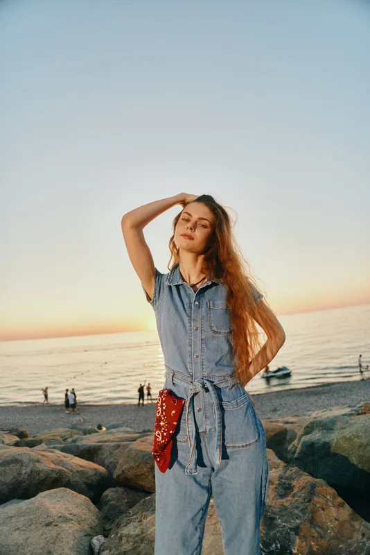 Lifestyle woman in modern boho-western denim street style posing on rocky beach at sunset with relaxed expression and film color tones enhancing warm atmosphere.