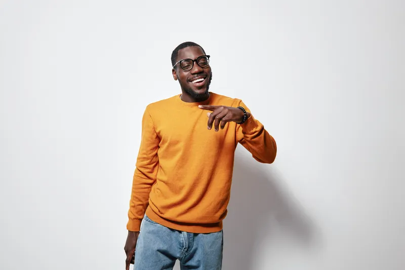 African American man with dark skin wearing orange sweater and glasses smiling and pointing while standing against white background in casual style.