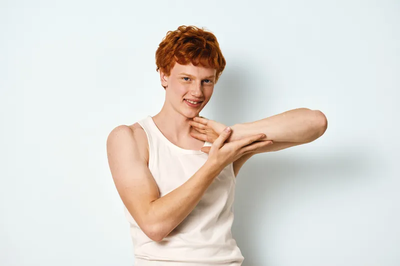 Young man with red hair in white sleeveless shirt stretching his arm and smiling against plain white background, casual portrait with relaxed body language and natural expression.
