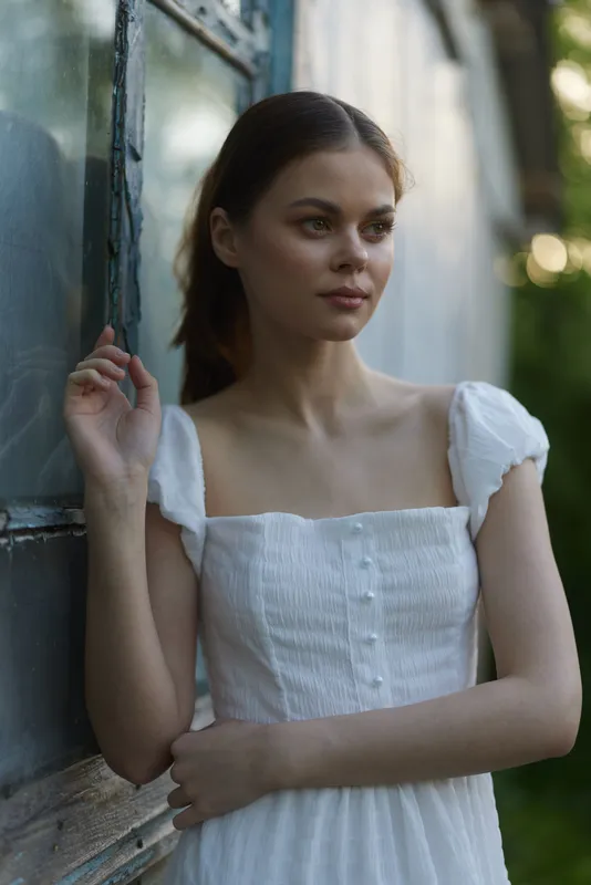 Young woman in a white dress by a vintage window, evoking a serene and thoughtful mood with soft natural light enhancing her beauty