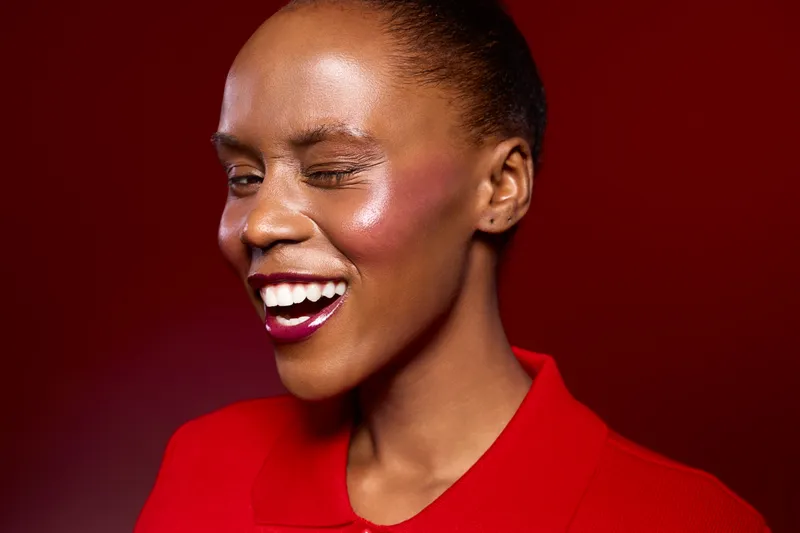Happy African American woman with dark skin wearing red shirt winks and smiles on red background. Close-up portrait with natural makeup and positive facial expression concept.