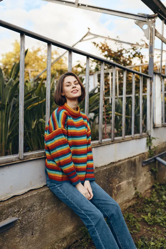 Young woman wearing a colorful striped sweater and casual jeans, sitting and leaning on a greenhouse railing, smiling in warm autumn light, relaxed lifestyle portrait for fashion and travel.