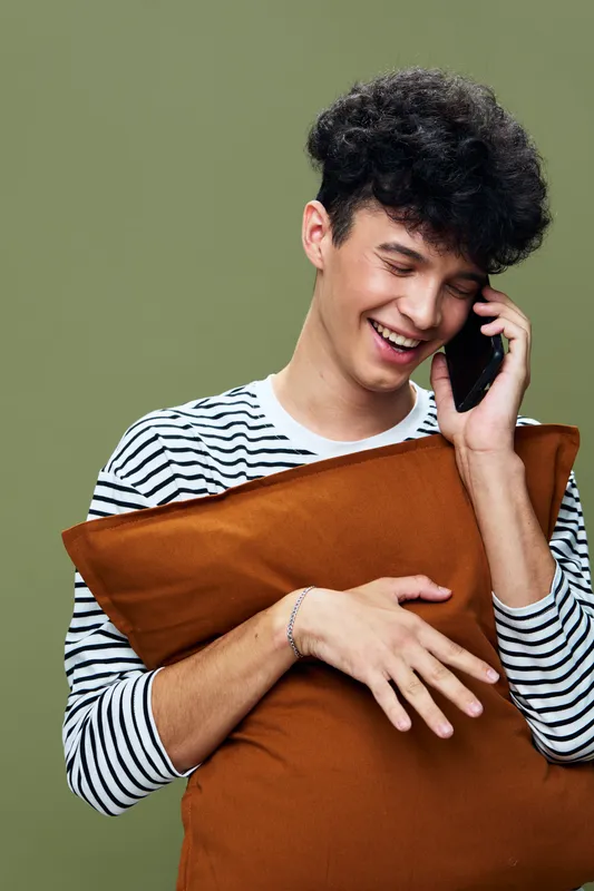 smiling young man talking phone holding brown pillow wearing striped shirt with curly hair on olive background, casual lifestyle happiness and communication concept
