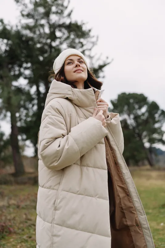 Woman wearing a beige puffer coat and beanie stands outdoors in a park, looking up with a calm expression, cozy winter fashion in a natural, spacious setting.