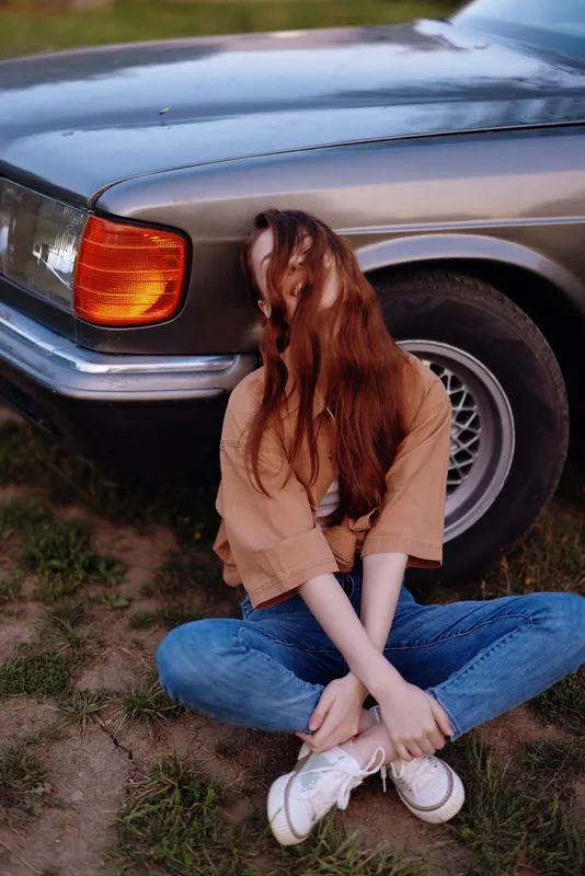 A young woman with long hair seated next to a vintage car, dressed casually in a brown shirt and blue jeans, exuding a relaxed and carefree vibe in a green outdoor setting.