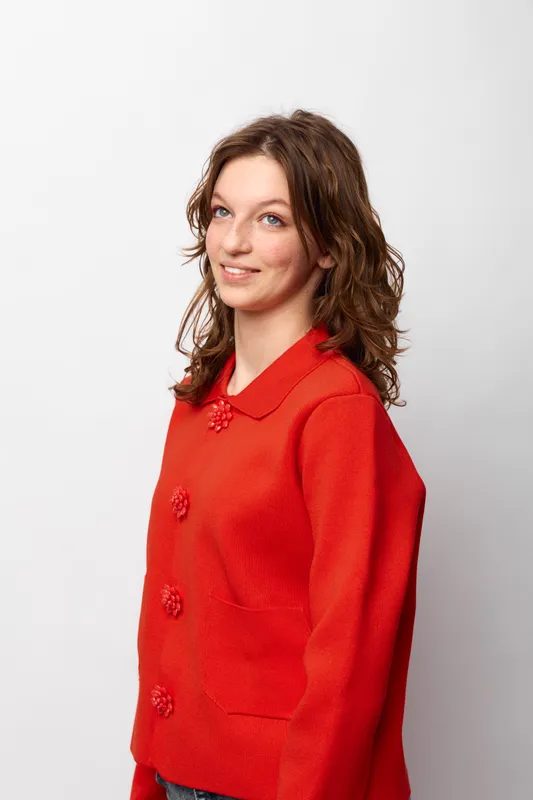 Young woman with curly hair wearing a red sweater smiles gently, posing against a white background. Casual style, natural look, and relaxed mood concept.
