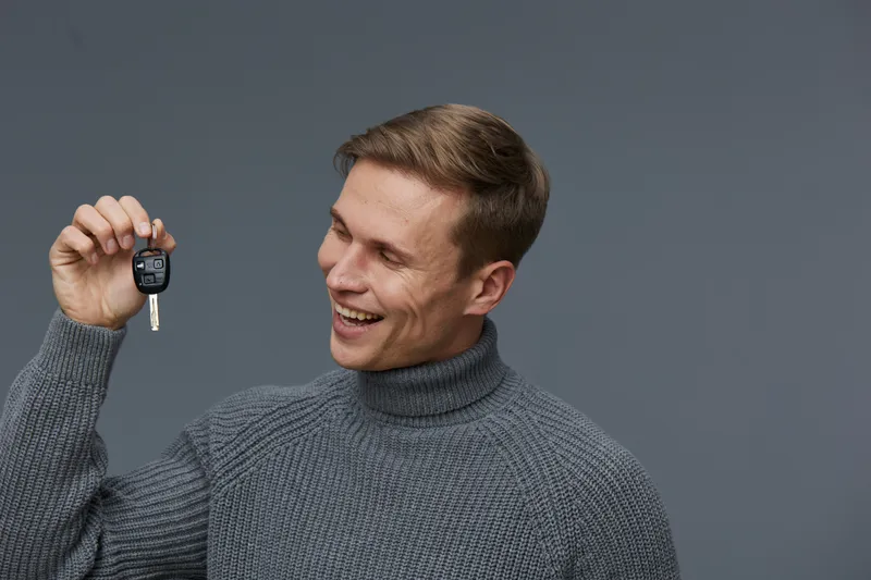 Smiling man holding car key with remote control in hand, wearing cozy gray turtleneck sweater, looking happy, isolated on plain solid gray background, studio portrait. People lifestyle concept