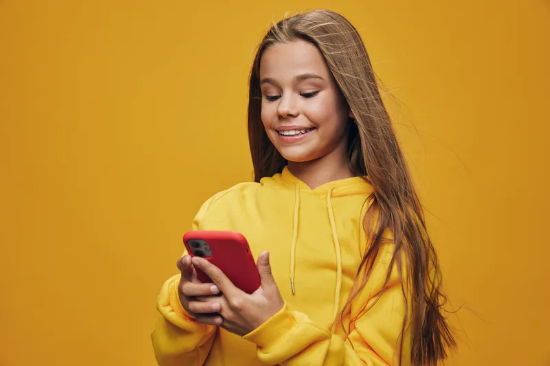 Girl smiling and using smartphone with red case, wearing yellow hoodie, standing against orange background while looking at screen for texting or social media.