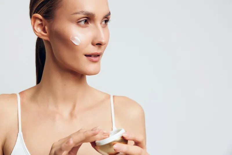 Young woman with long hair applies white face cream on her cheek while holding a jar in a bright studio with a plain background expressing skincare and self-care routine.