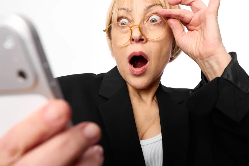 Elderly woman with glasses shocked by smartphone, hand near face, surprised astonished expression for online communication, bright studio indoors moment.