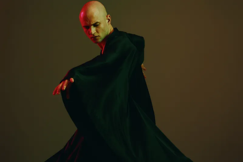 A man in a dark robe gracefully moves in a captivating dance, emphasizing the fluidity of motion. The image features a minimalist background with muted tones, highlighting the elegance of the moment.