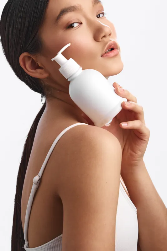 Asian woman holds a skincare bottle in a studio, pump pressed gently to her skin, soft light and clean backdrop create a calm, modern beauty atmosphere.